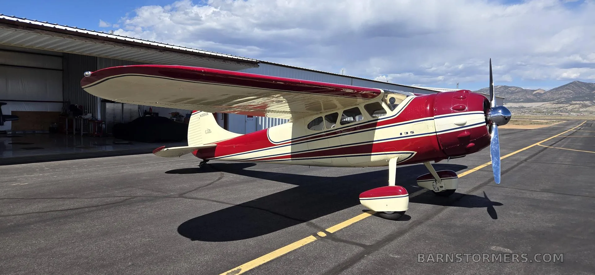 A red and cream vintage single-engine propeller aircraft parked on an asphalt tarmac next to a hangar.