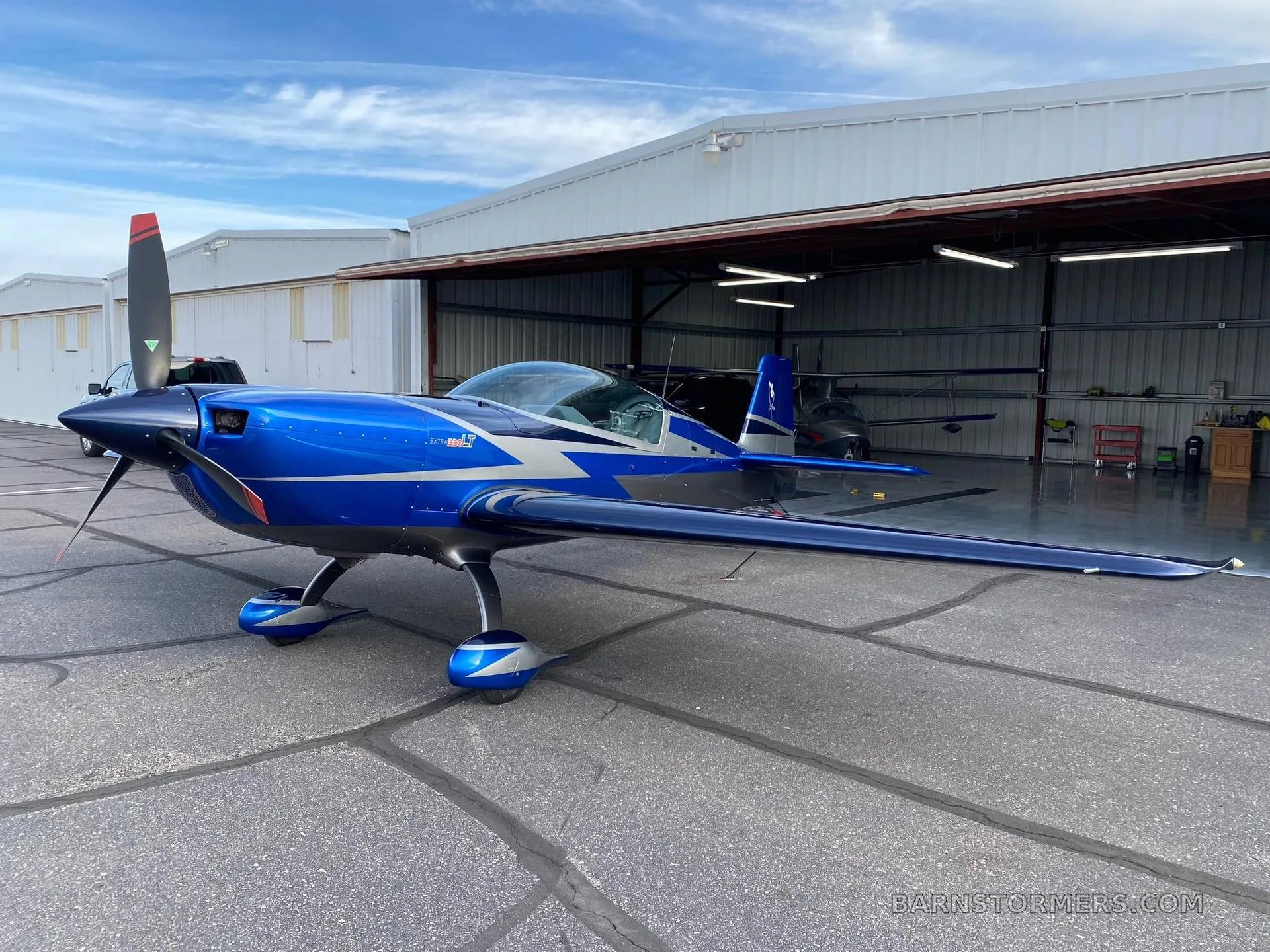 A blue and silver Extra 300LT aerobatic aircraft is parked on an asphalt apron in front of a hangar under a partly cloudy sky.