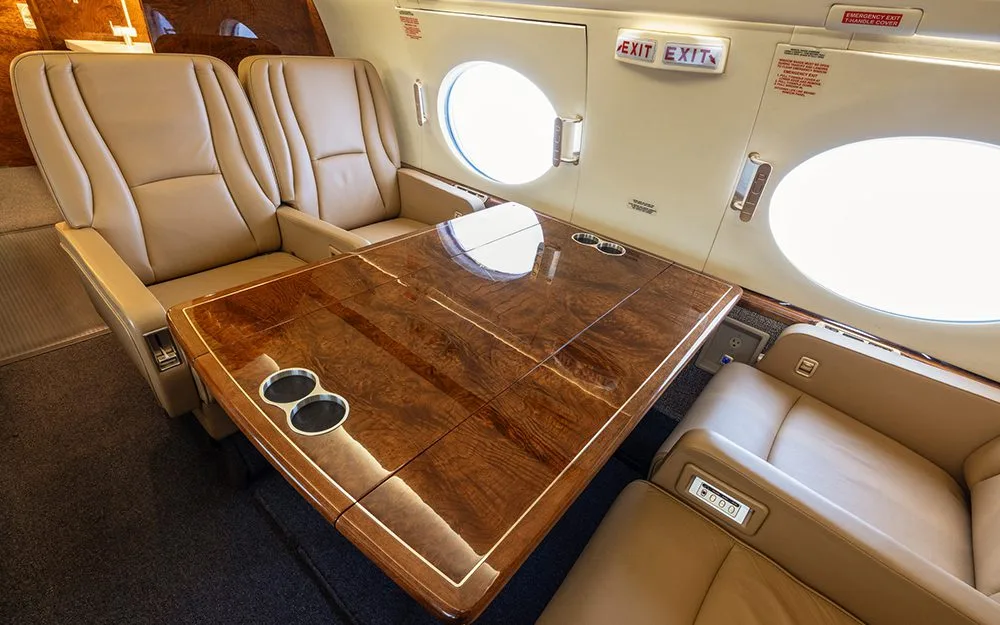 A close-up view of a polished wood table and beige leather seats in a private jet cabin.
