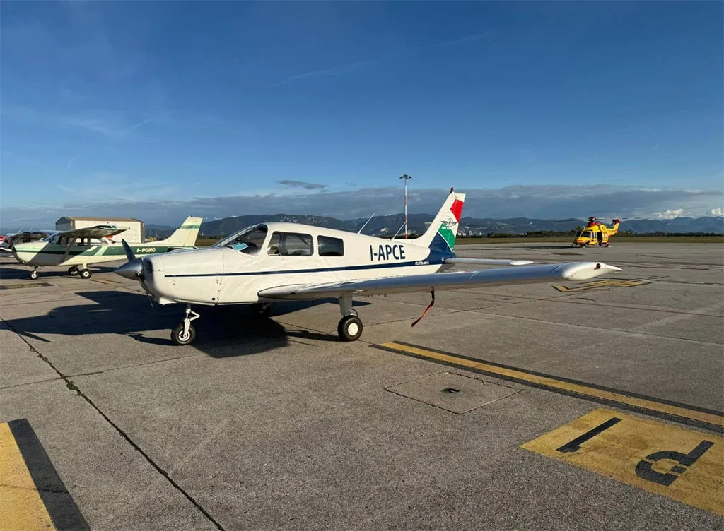 A white single-engine propeller aircraft with "I-APCE" registration parked on an airfield with other aircraft in the background.