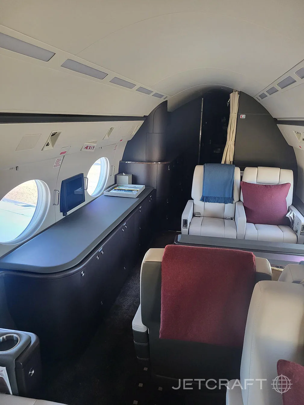 A rear view of the business jet cabin showing a dark credenza along the left wall and two light grey seats in the background.