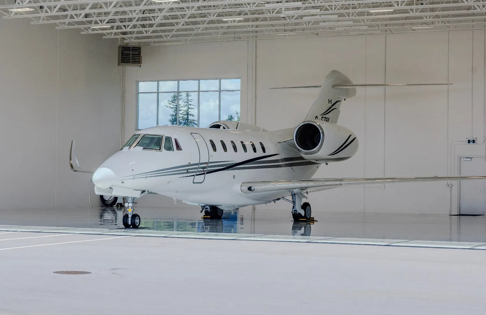A white business jet with dark grey stripes is parked inside a bright, modern hangar, viewed from the front left.