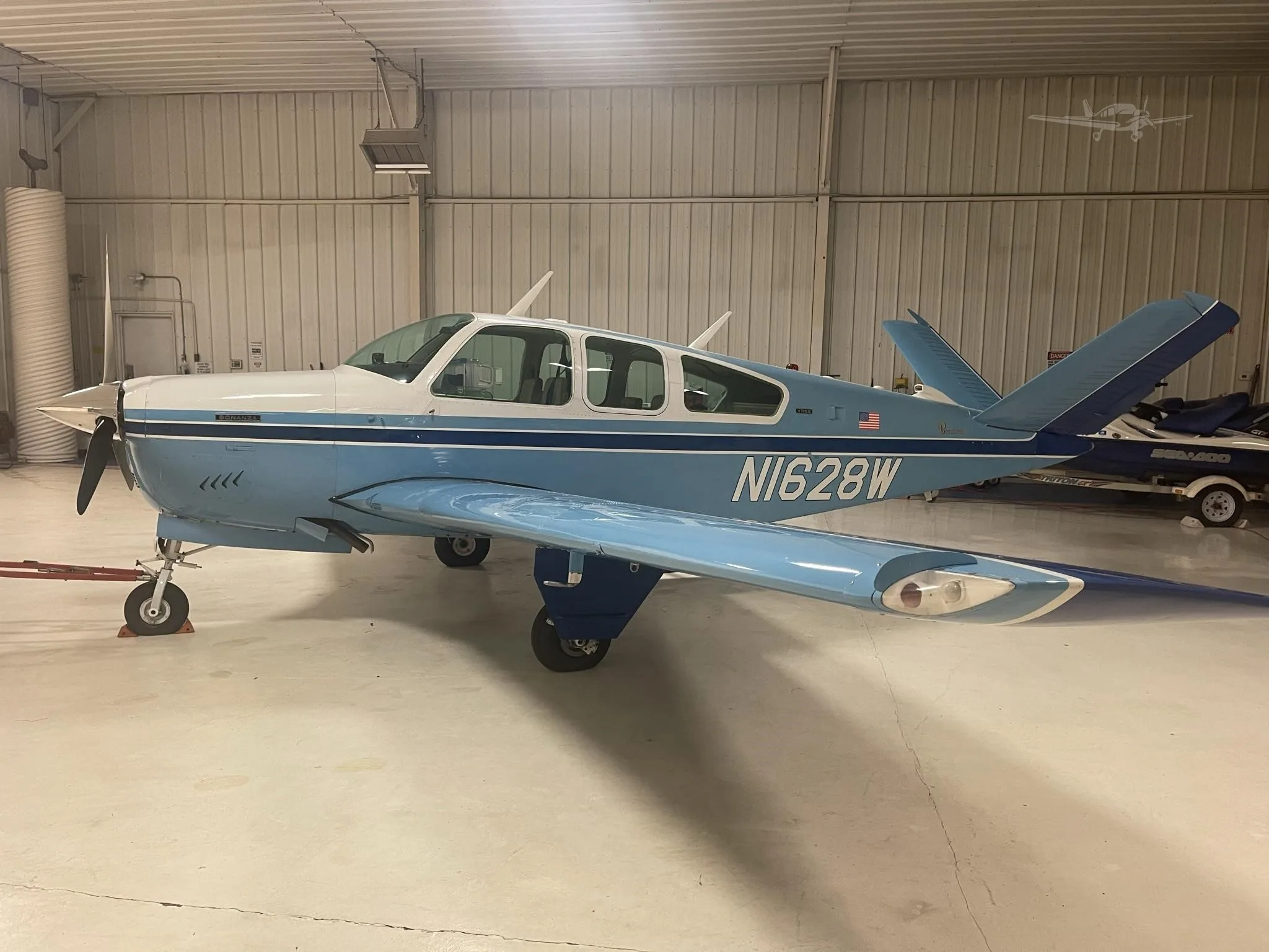 A light blue and white Beechcraft Bonanza with dark blue stripes, registration N1628W, is parked inside a hangar.