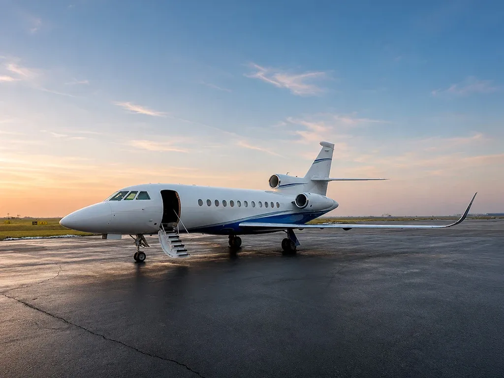 A white and blue Dassault Falcon business jet with its door open and stairs deployed, parked on a wet tarmac at sunset.