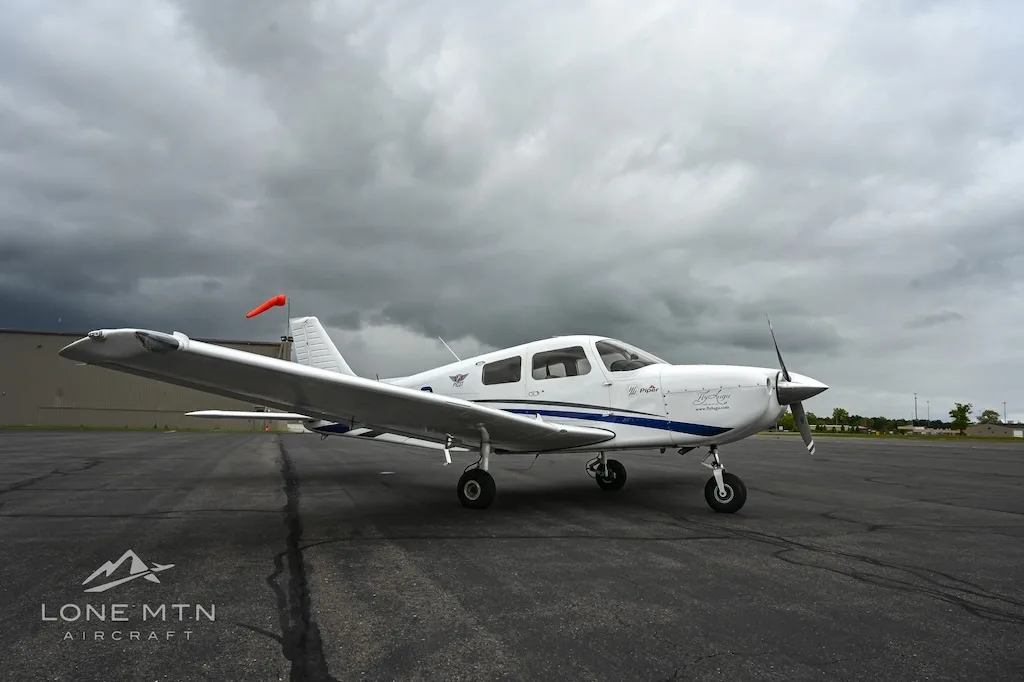 A white single-engine propeller aircraft with blue accents parked on an asphalt tarmac under a cloudy sky.