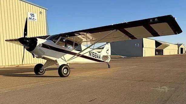 A white and black high-wing single-engine aircraft, N168AA, is parked on an airfield with hangars in the background.