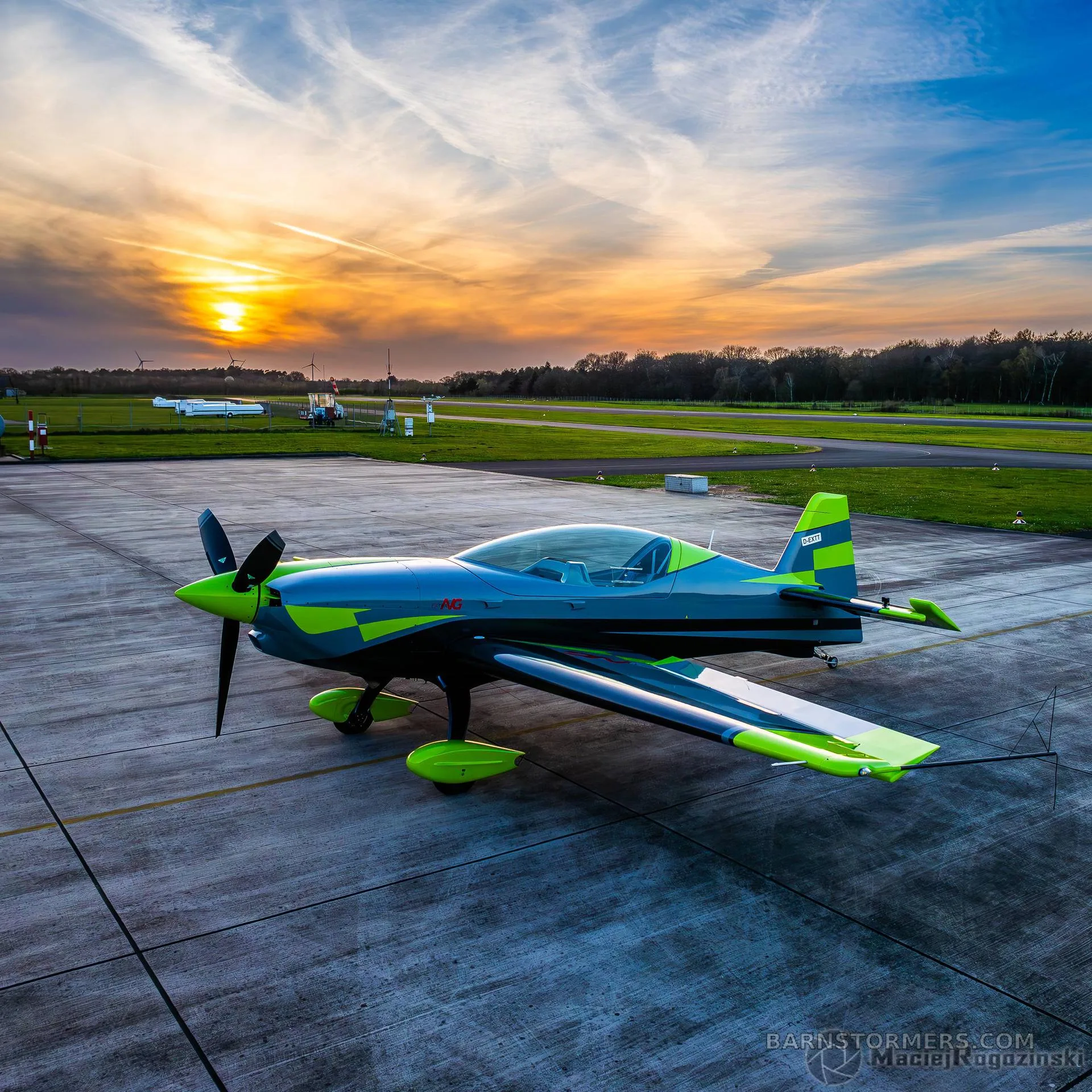 A grey and neon green Extra 330SC aircraft is parked on an airfield at sunset.