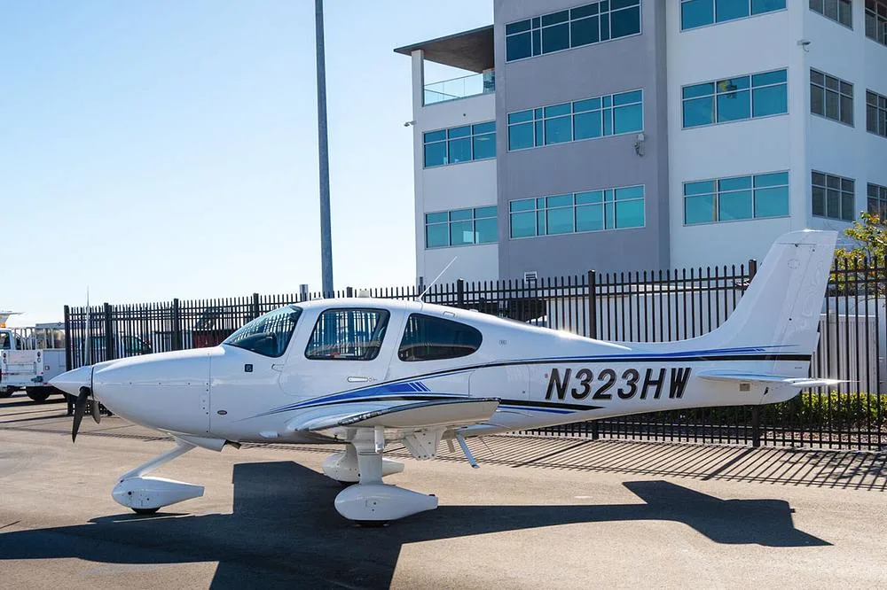 A white Cirrus SR22 aircraft with blue and black stripes, registration N323HW, parked on an asphalt ramp next to a modern building.