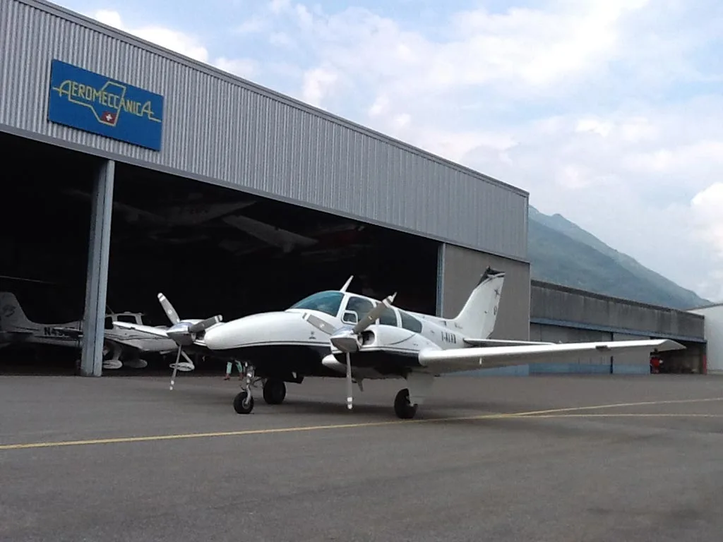 A white and black twin-engine aircraft parked on an tarmac in front of a hangar with "Aeromeccanica" sign and mountains in the background.
