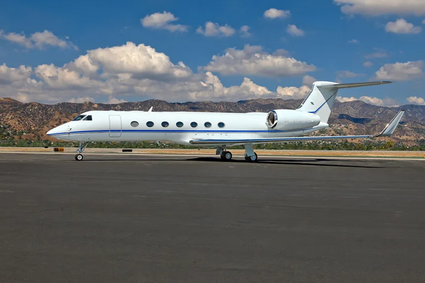 A white Gulfstream business jet with a blue stripe is parked on an asphalt runway with mountains and a blue sky with clouds in the background.