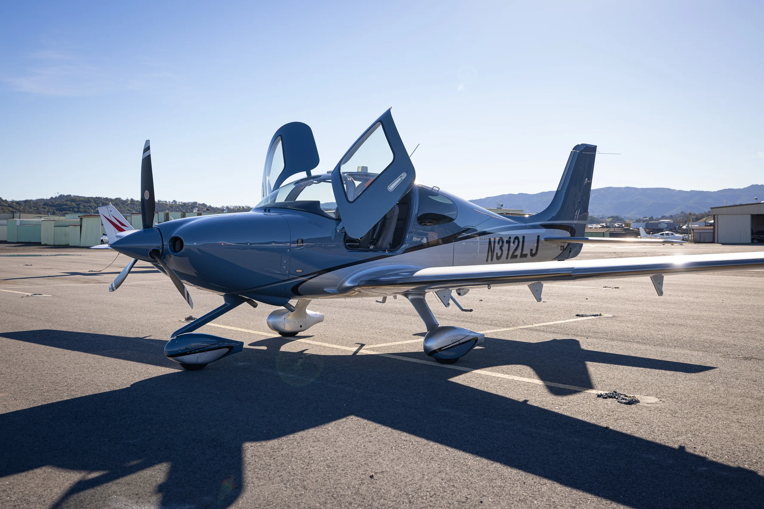 A blue and silver single-engine propeller aircraft with its gull-wing doors open, parked on an airport apron under a clear sky.