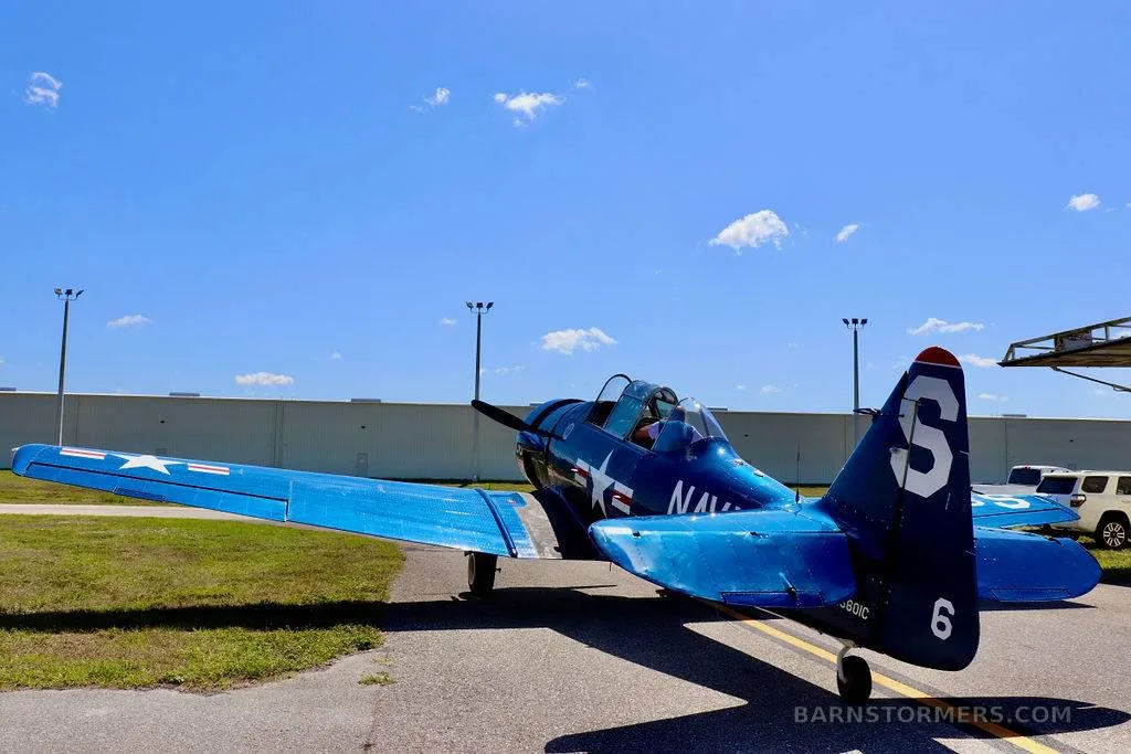 A dark blue Vought F4U Corsair aircraft with a distinctive gull-wing design is parked on an airfield, viewed from the rear.
