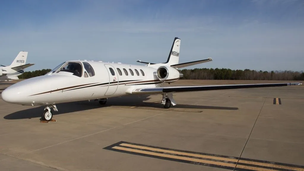 A white Cessna Citation CJ3 private jet with black and brown stripes, tail number N556MW, parked on an airport tarmac under a clear blue sky.