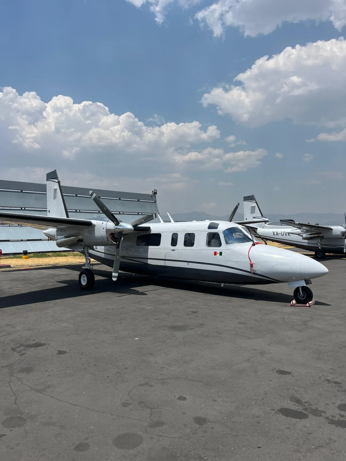A white and dark grey twin-engine turboprop aircraft with a Mexican flag on the nose, parked on an airport tarmac with other aircraft in the background.