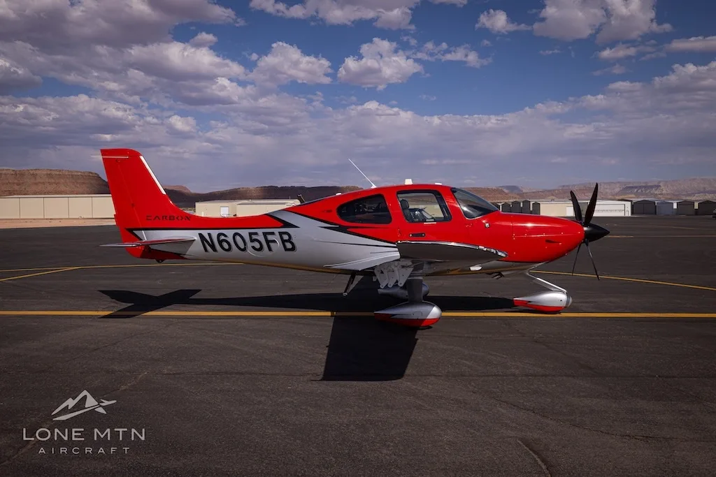 A side profile view of a red and silver Cirrus SR22T aircraft (N605FB) on an airfield under a partly cloudy sky.