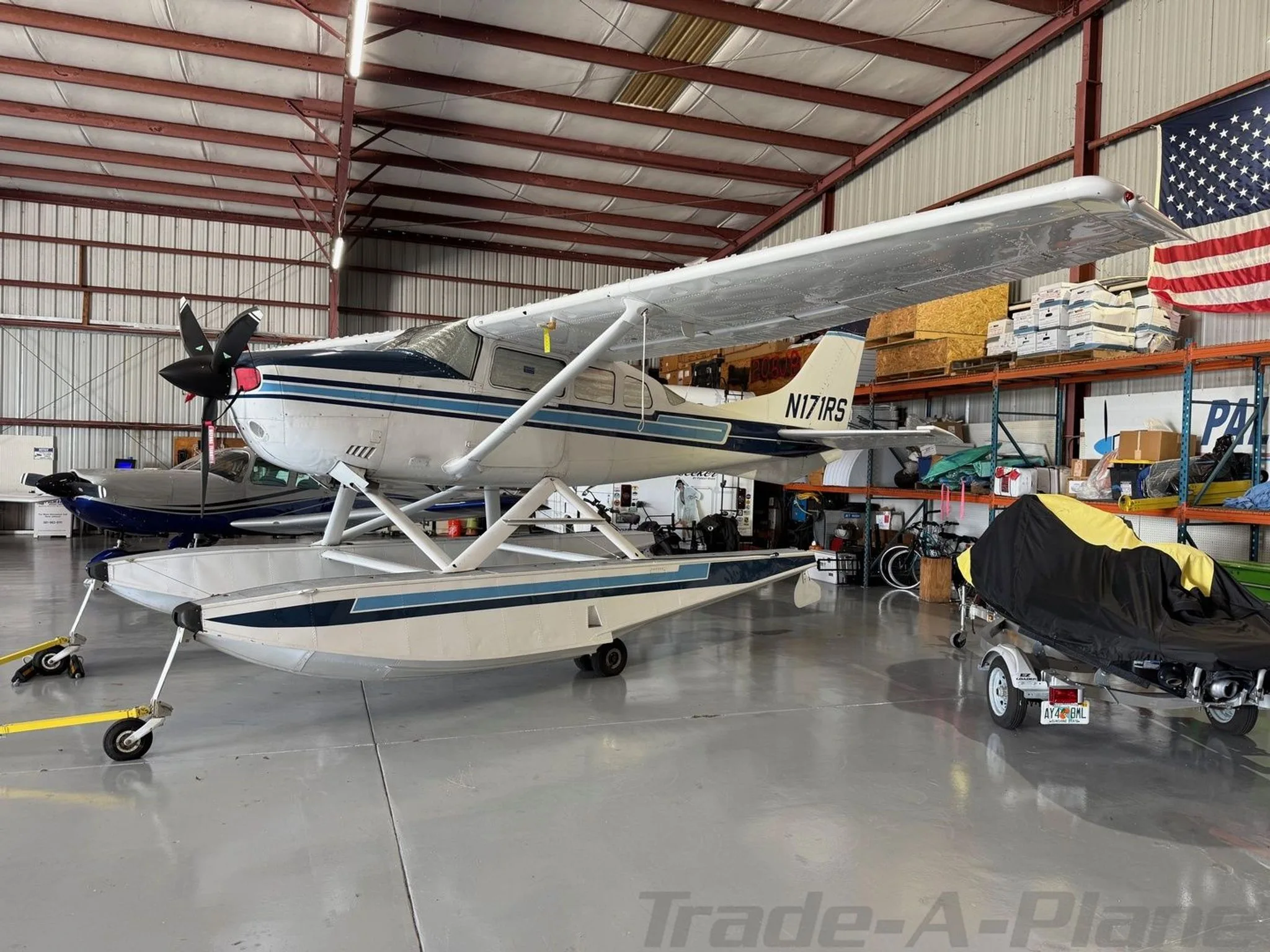 Side view of a white and blue Cessna 206 Stationair floatplane, registration N171RS, parked in a hangar.