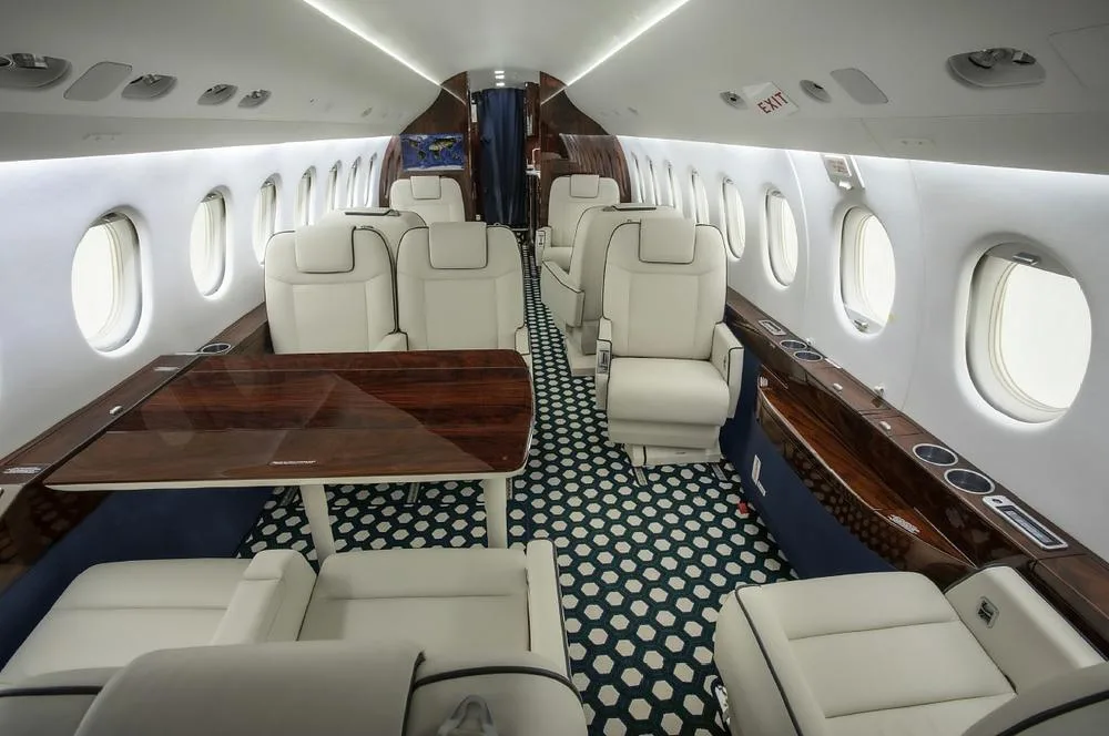 The spacious interior cabin of a business jet featuring white seats, a dark wood table, and patterned carpet.