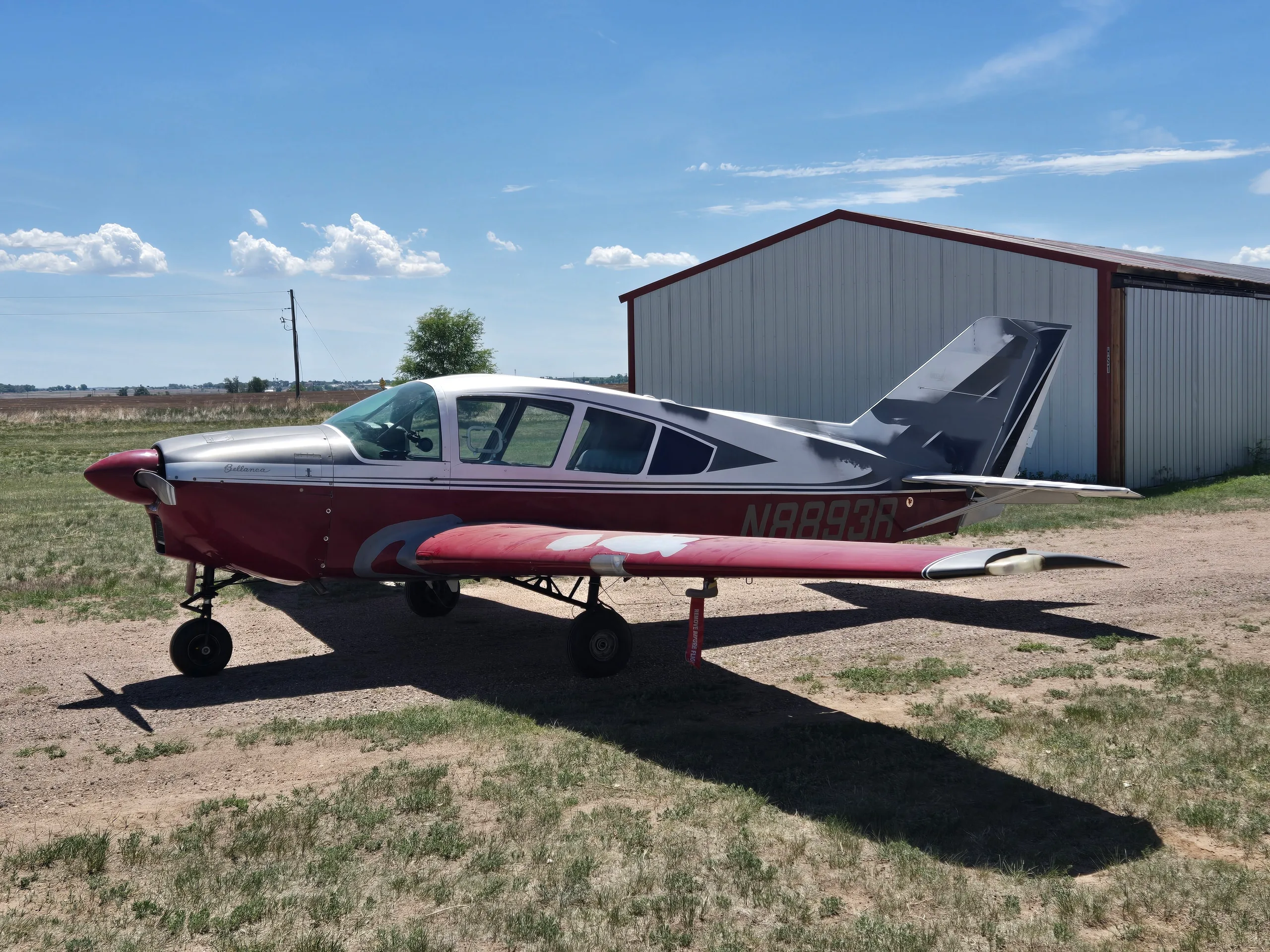 A red and silver single-engine aircraft, registration N8893R, parked on a dirt lot next to a white hangar under a blue sky.