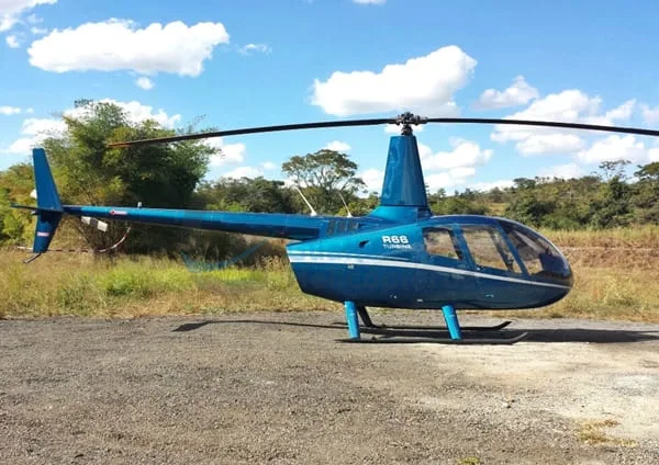 A blue Robinson R66 Turbine helicopter with white stripes sits on a gravel patch under a partly cloudy sky.