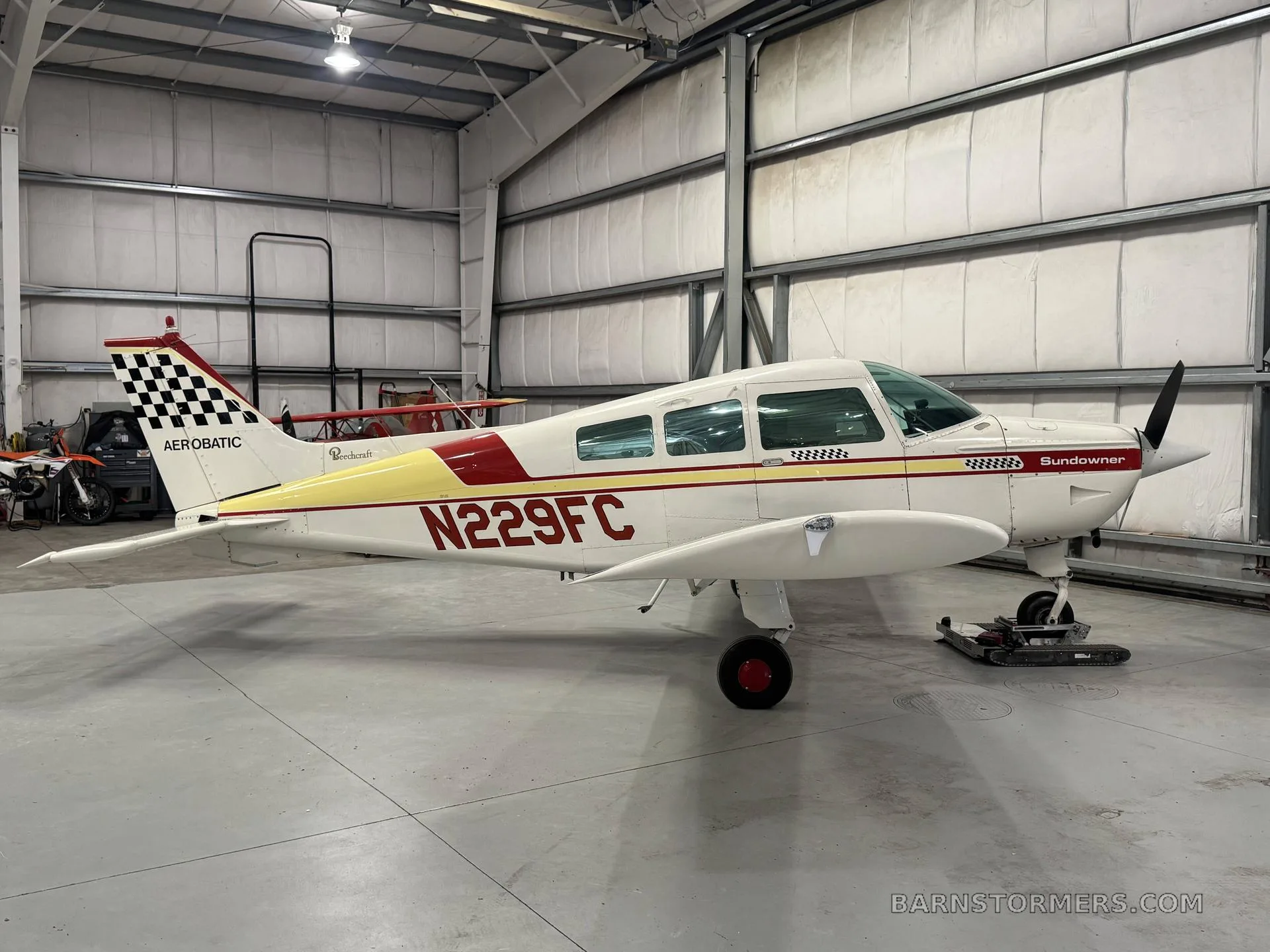 A white and red Beechcraft Sundowner N229FC with a checkered tail is parked in a hangar, viewed from the side.