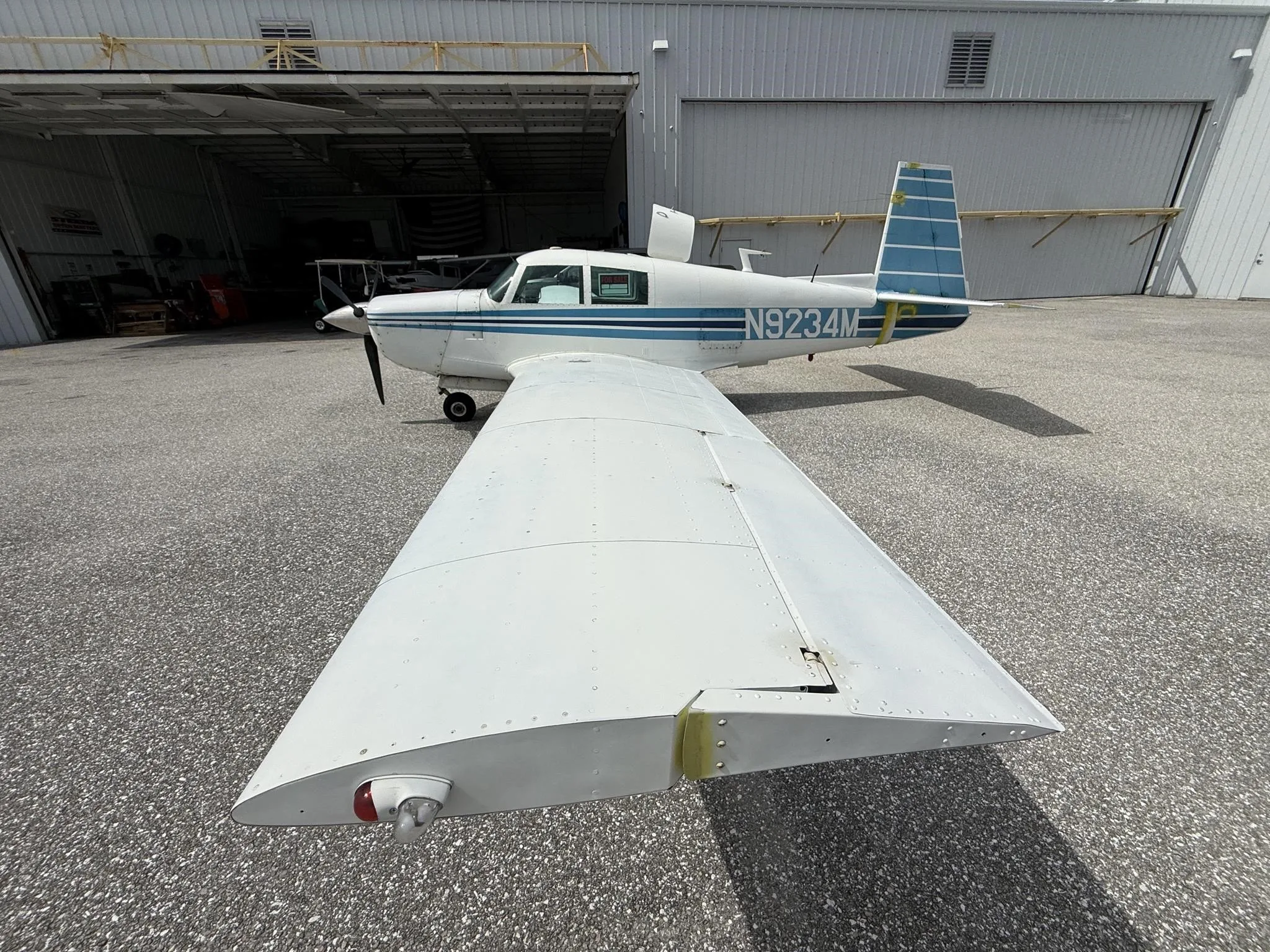 A white single-engine aircraft with blue stripes and registration N9234M, parked on an asphalt apron in front of an open hangar.