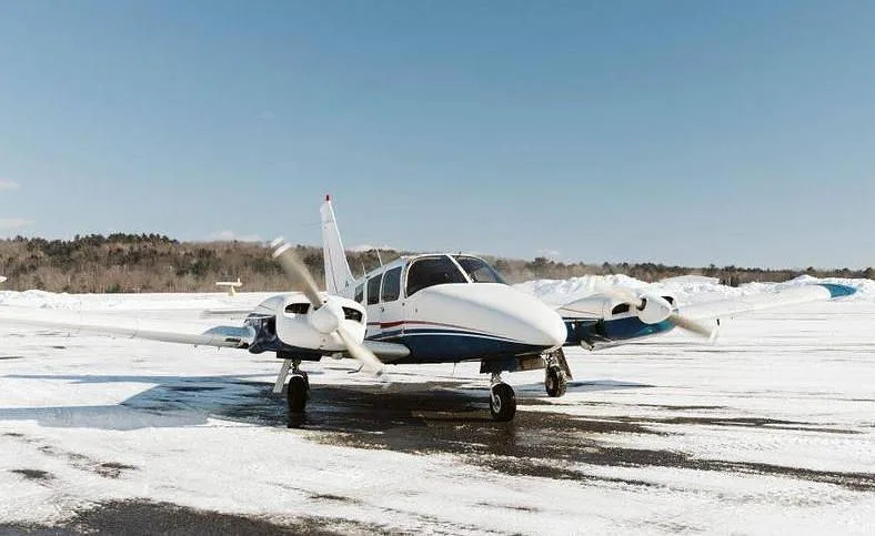A white and blue twin-engine Piper Seneca I aircraft is parked on a snowy tarmac with trees in the background.