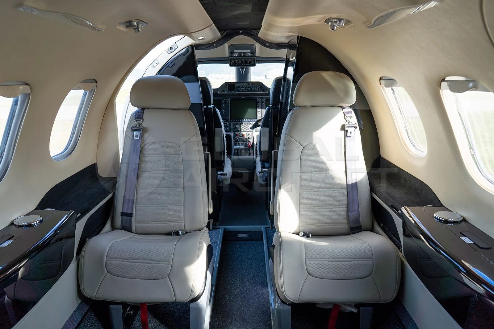 The interior cabin of an Embraer Phenom 100 shows light beige leather seats and a view into the cockpit.
