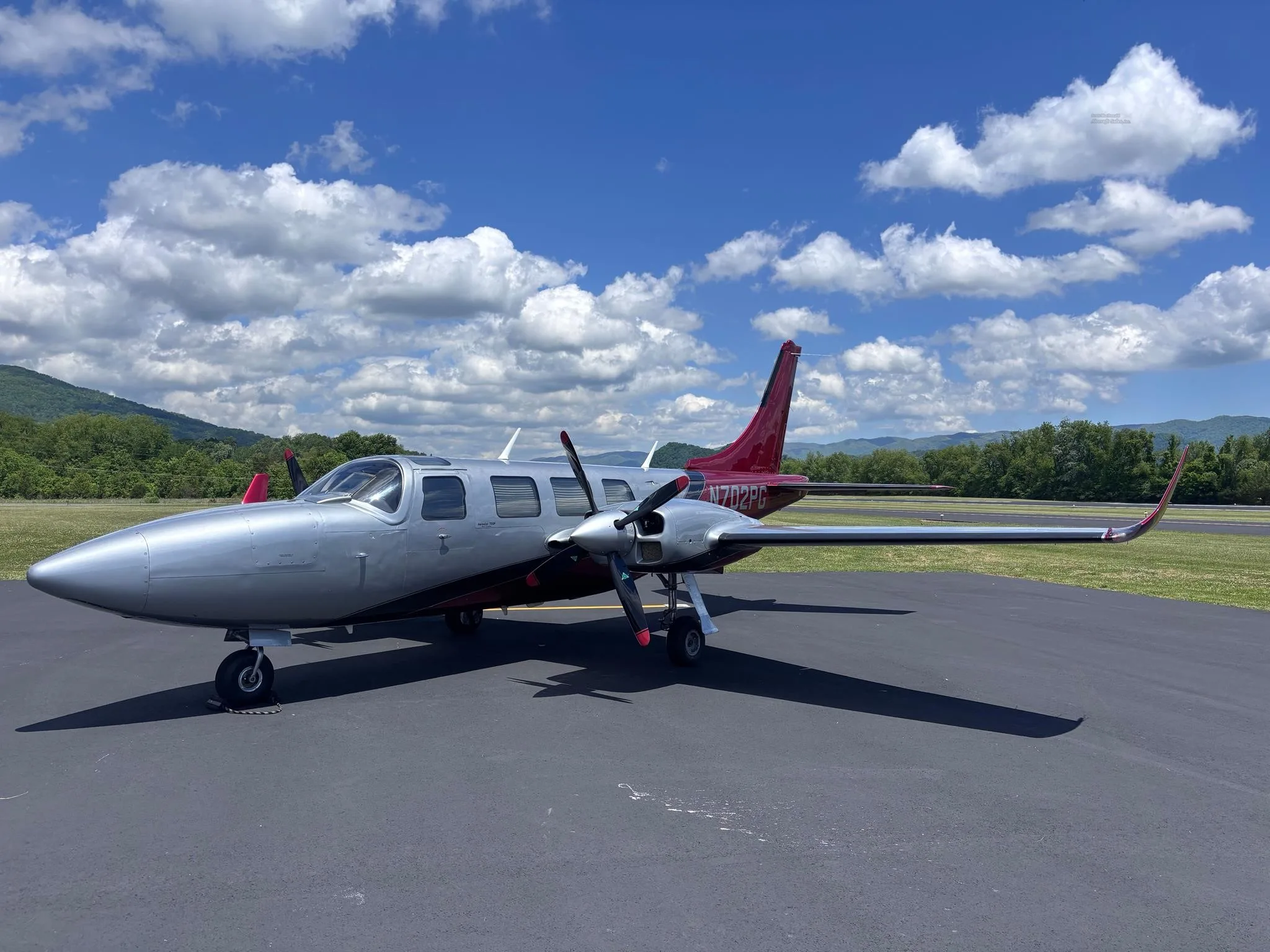 A silver twin-engine propeller aircraft with a red tail (N702PG) is parked on an asphalt tarmac under a blue sky with white clouds, with green mountains in the background.