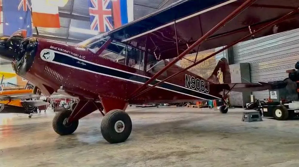 A burgundy Aviat Aircraft Husky A-1C, registration N90BJ, with black and white stripes, parked in a hangar.