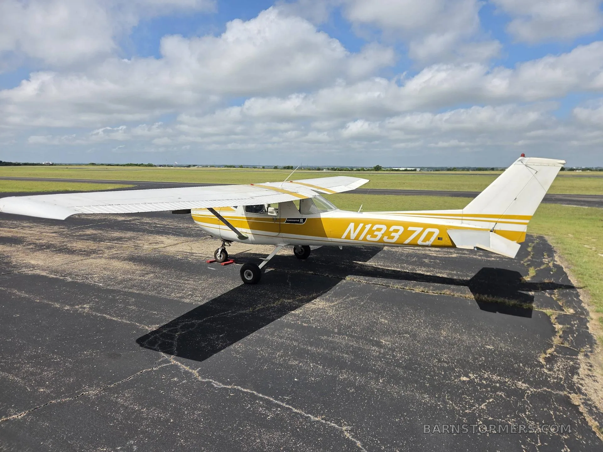 A white and yellow light aircraft (N13370) with a high wing, parked on an asphalt ramp, viewed from the rear-left.