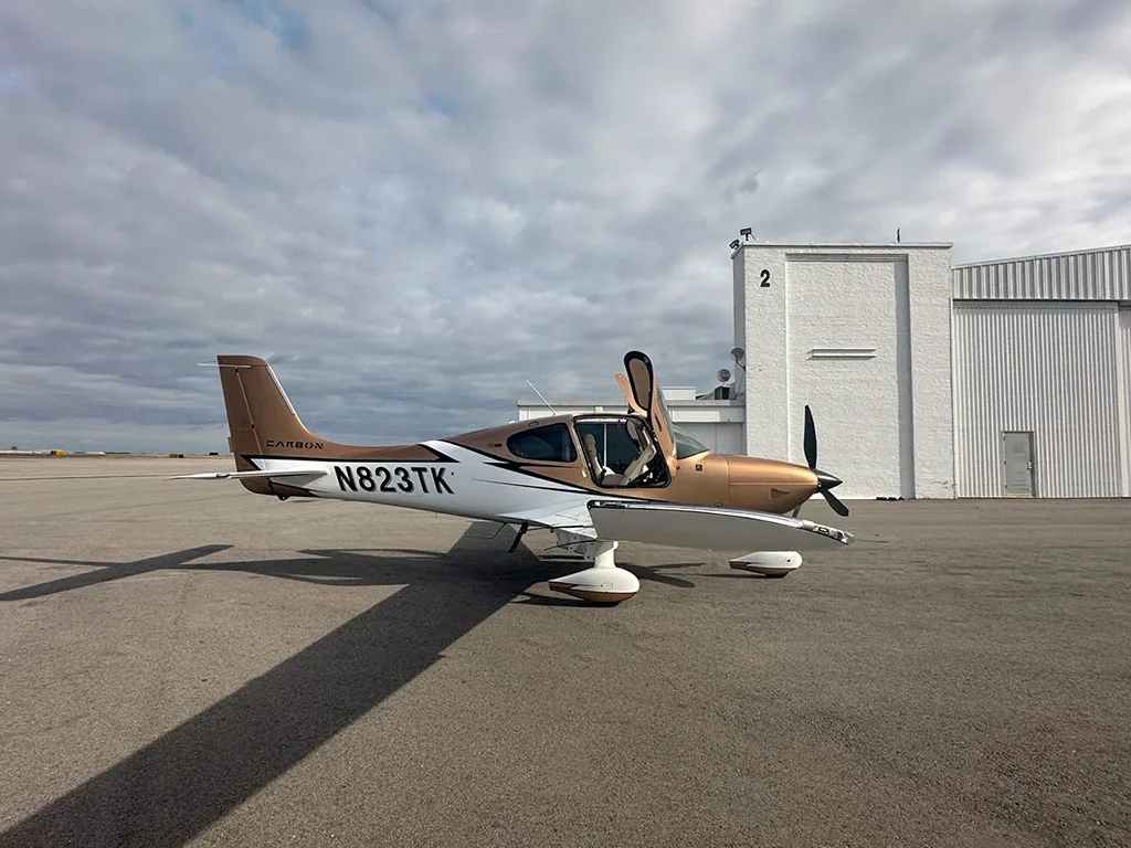 A Cirrus SR22 aircraft, N823TK, with its doors open, parked on an airfield next to a white building.