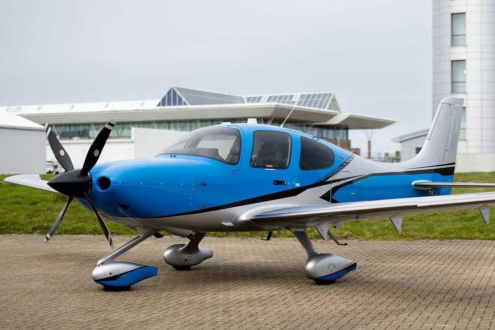 A blue and silver Cirrus SR22T GTS aircraft parked on a tarmac with modern buildings in the background.