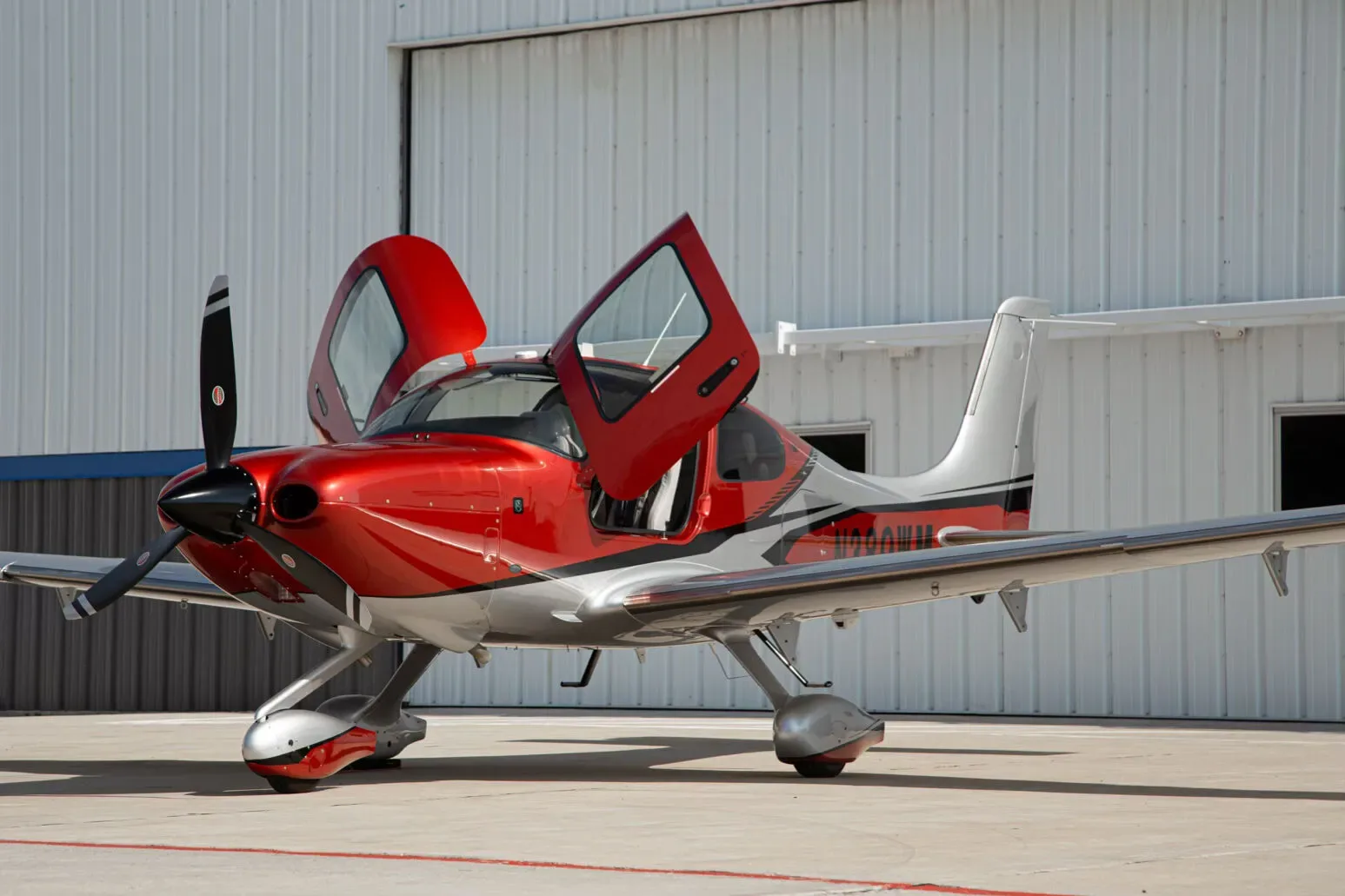 A red and silver single-engine propeller aircraft, N389WM, with its doors open, parked on an airfield in front of a white hangar.