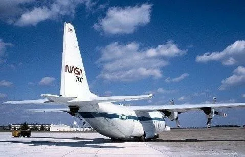 A white NASA C-130 aircraft with blue stripes is parked on an airfield tarmac.