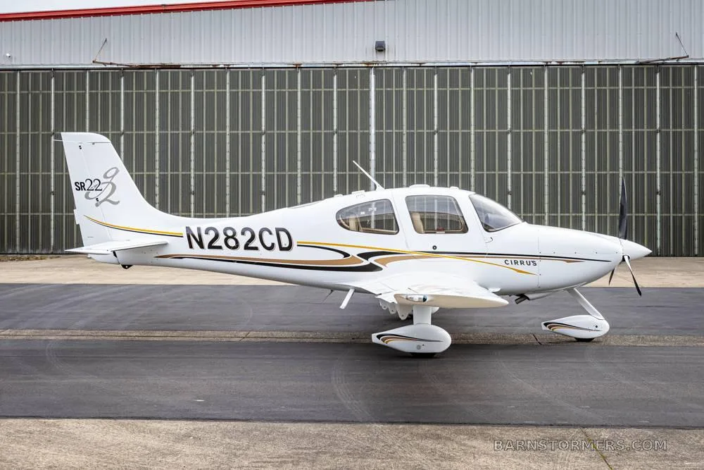 A white Cirrus SR22 aircraft with black and gold stripes is parked on an asphalt tarmac in front of a hangar, viewed from the front-right.