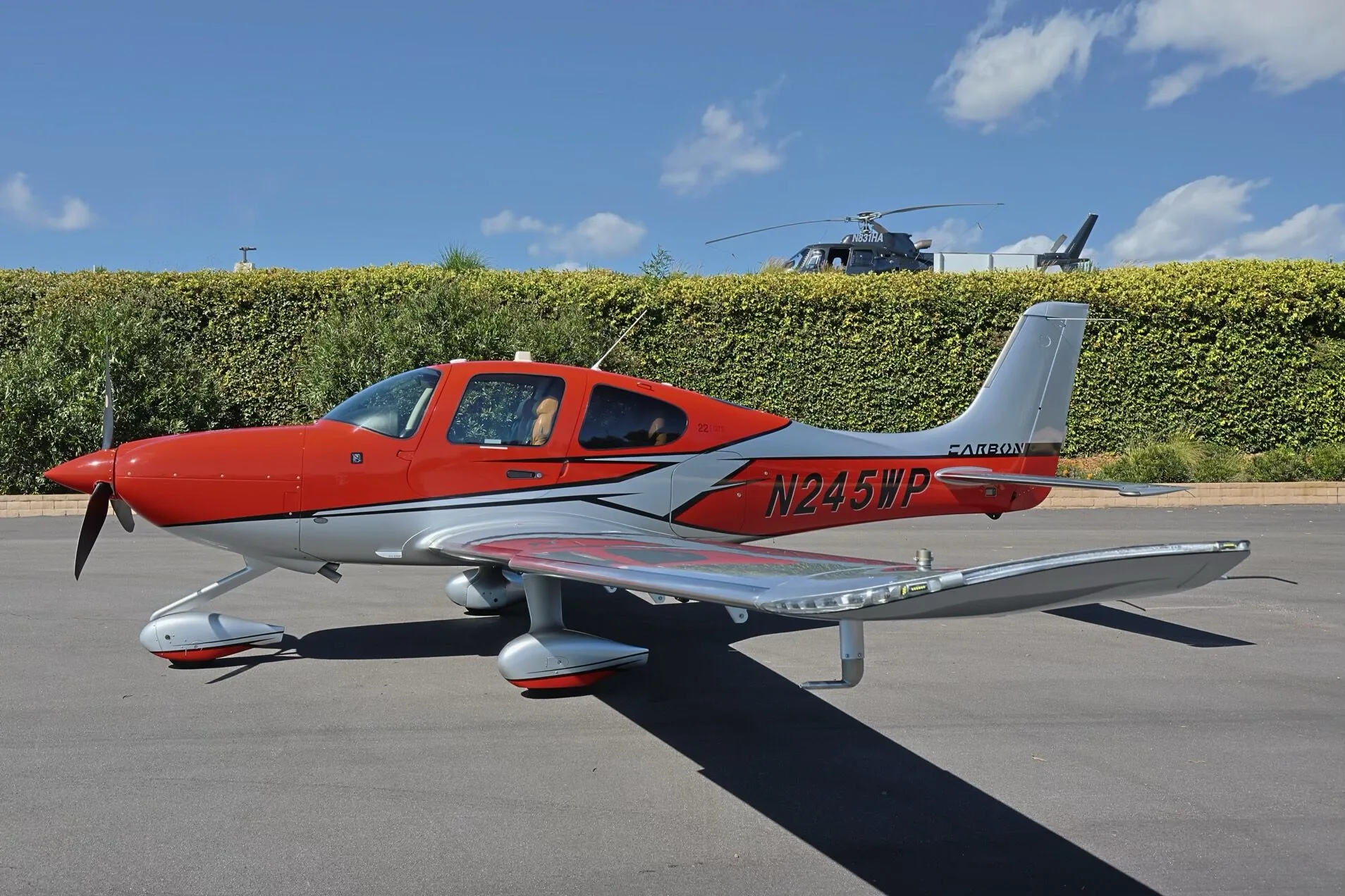 A red and silver Cirrus SR22 GTS aircraft, N245WP, parked on an tarmac with a helicopter partially visible.