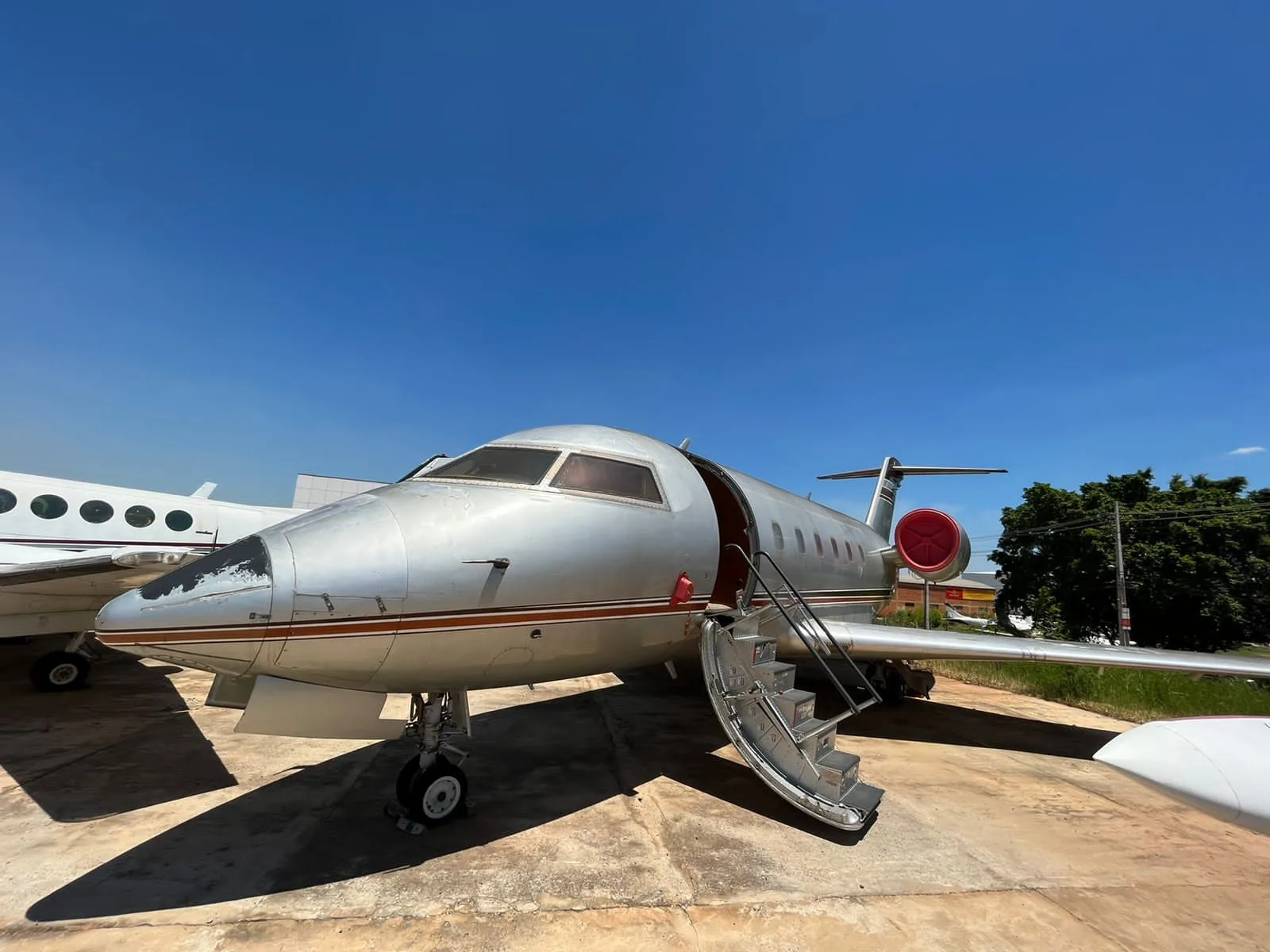A silver business jet with a red engine cover and open airstairs sits on a concrete tarmac under a clear blue sky.
