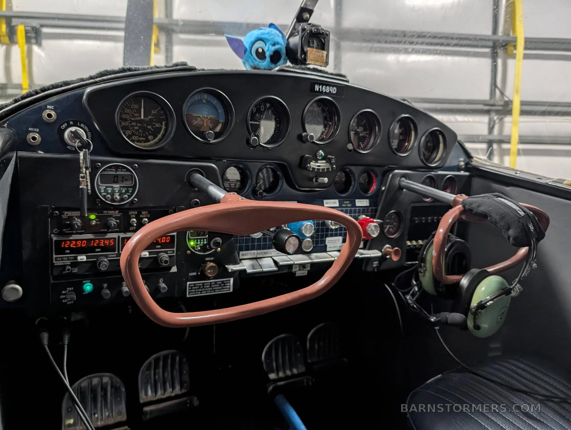 The cockpit of a light aircraft features an instrument panel with gauges, controls, and a small blue plush toy.