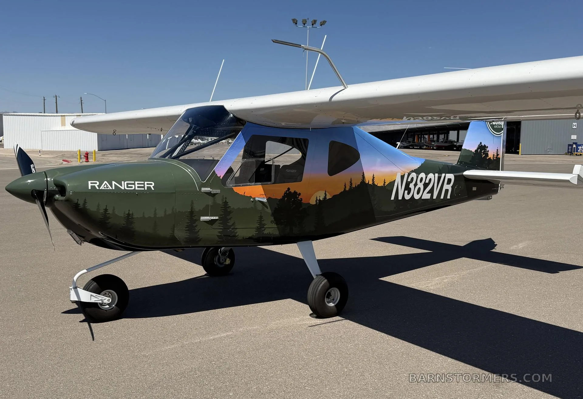A green and blue 'Ranger' light-sport aircraft with a forest and sunset paint scheme, seen from the front-right on an airfield.