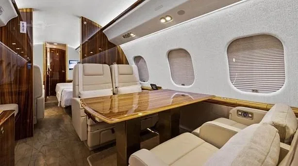 A luxurious aircraft interior features cream leather seats, a polished wooden table, and a private sleeping area in the background.