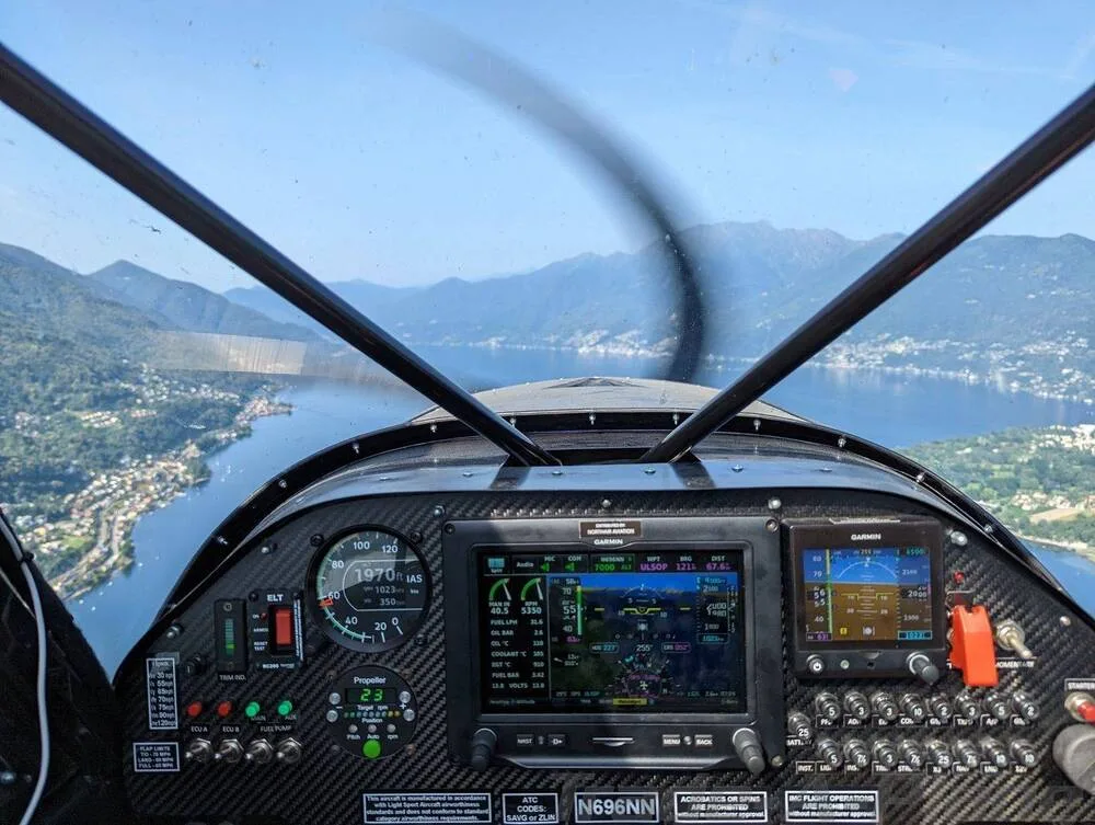 The cockpit view of a Zlin Shock Cub, showing the instrument panel with a Garmin display and a scenic view of a lake and mountains outside.