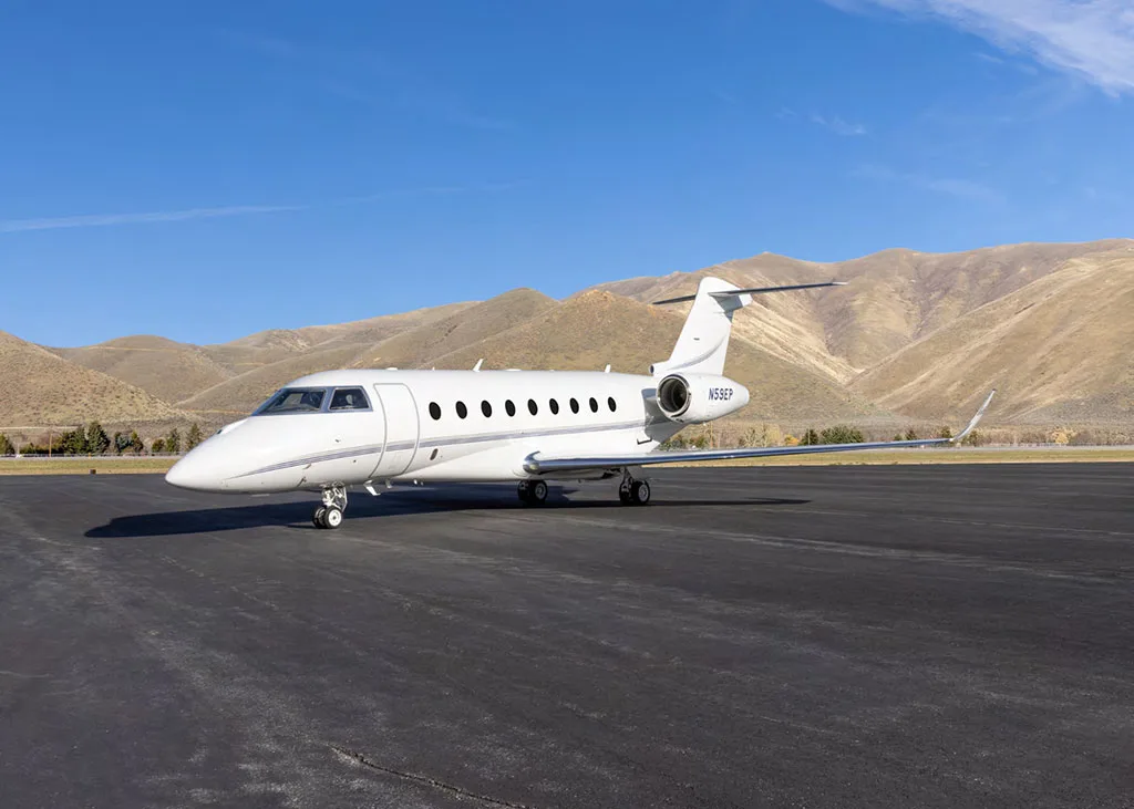 A white Gulfstream G550 business jet, tail number N59EP, is parked on an airfield with mountains in the background.