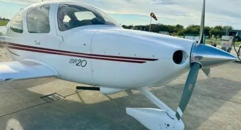 A white Cirrus SR20 aircraft with a red stripe on its fuselage, viewed from the front-right on a tarmac.