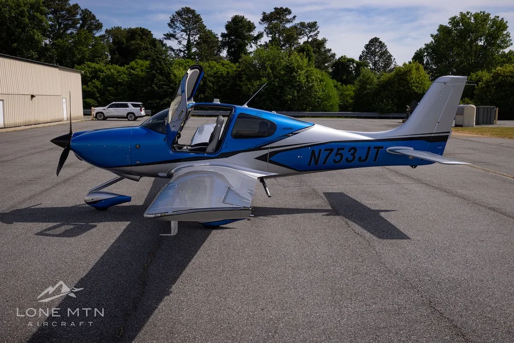 A blue and silver single-engine aircraft, N753JT, with open gull-wing doors is parked on an asphalt tarmac with trees in the background.