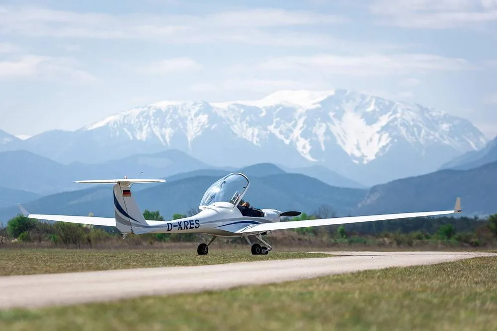 A white Diamond DA20 Katana aircraft with registration D-XRES is seen on a runway with snowy mountains in the background.
