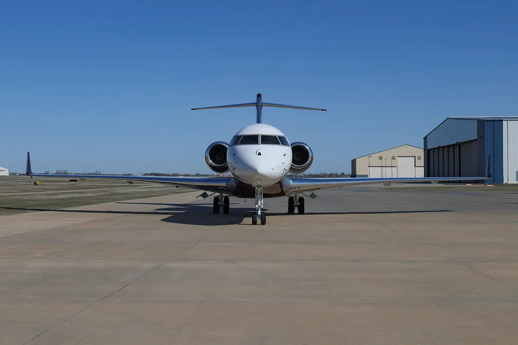 A Bombardier Global Express business jet with a white and maroon paint scheme, registration N919EM, viewed head-on on a tarmac with hangars in the background.