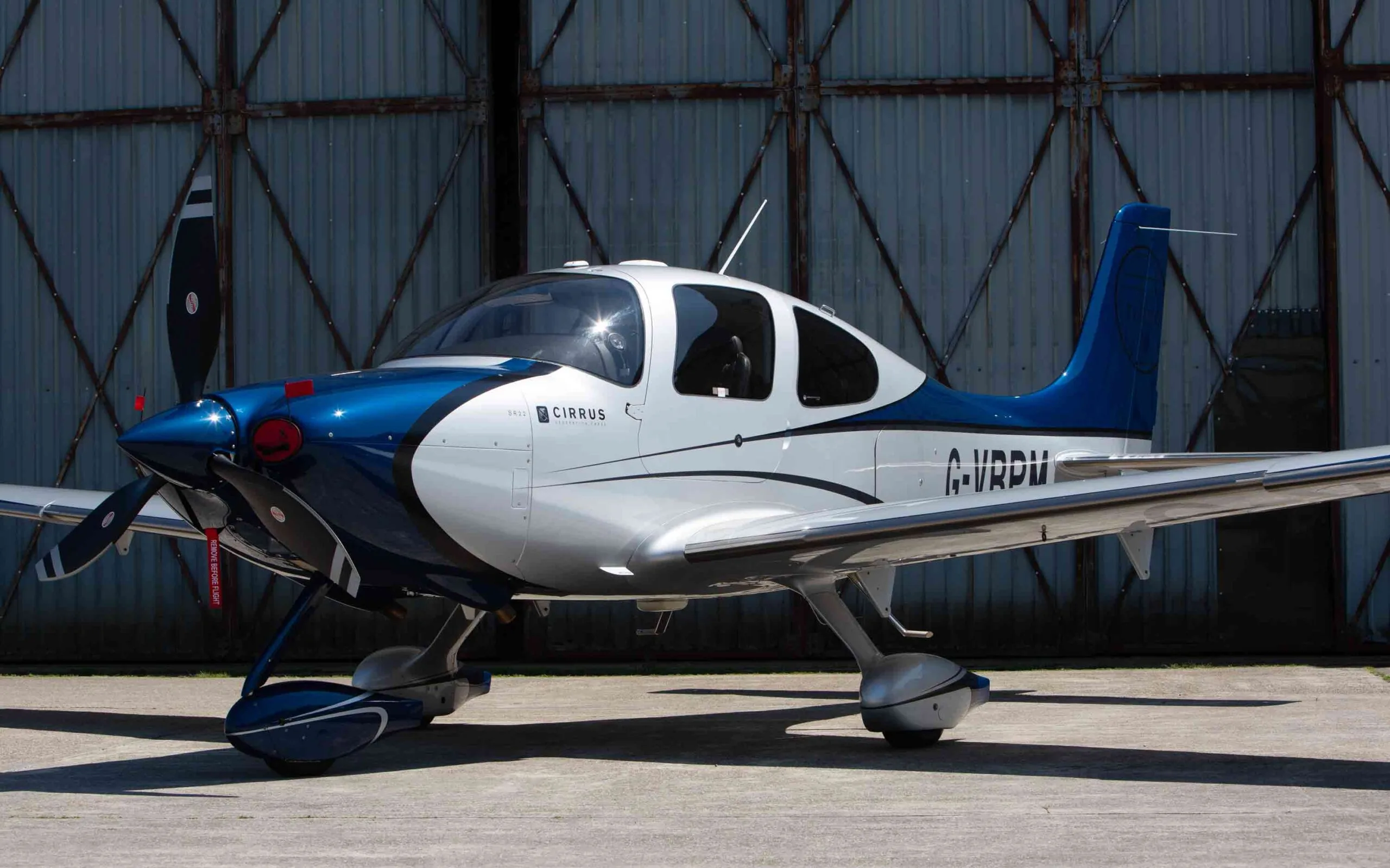 A side view of a blue and white Cirrus SR22 aircraft with registration G-VBPM, parked in front of a hangar.