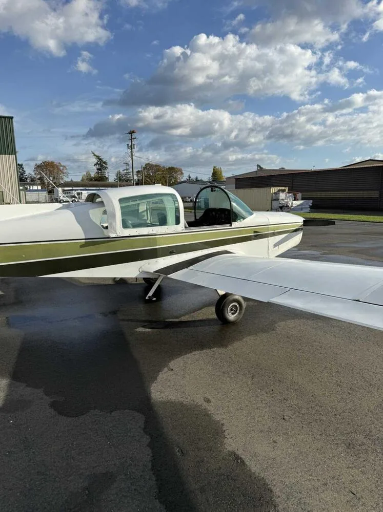 Side view of a white and green Grumman American AA-5 Traveler aircraft parked on an airfield under a partly cloudy sky.