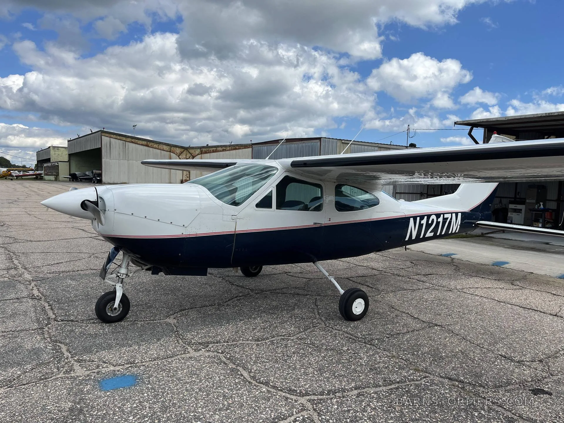 Three-quarter front view of the white and dark blue single-engine propeller aircraft N1217M, parked on a concrete apron under a partly cloudy sky.