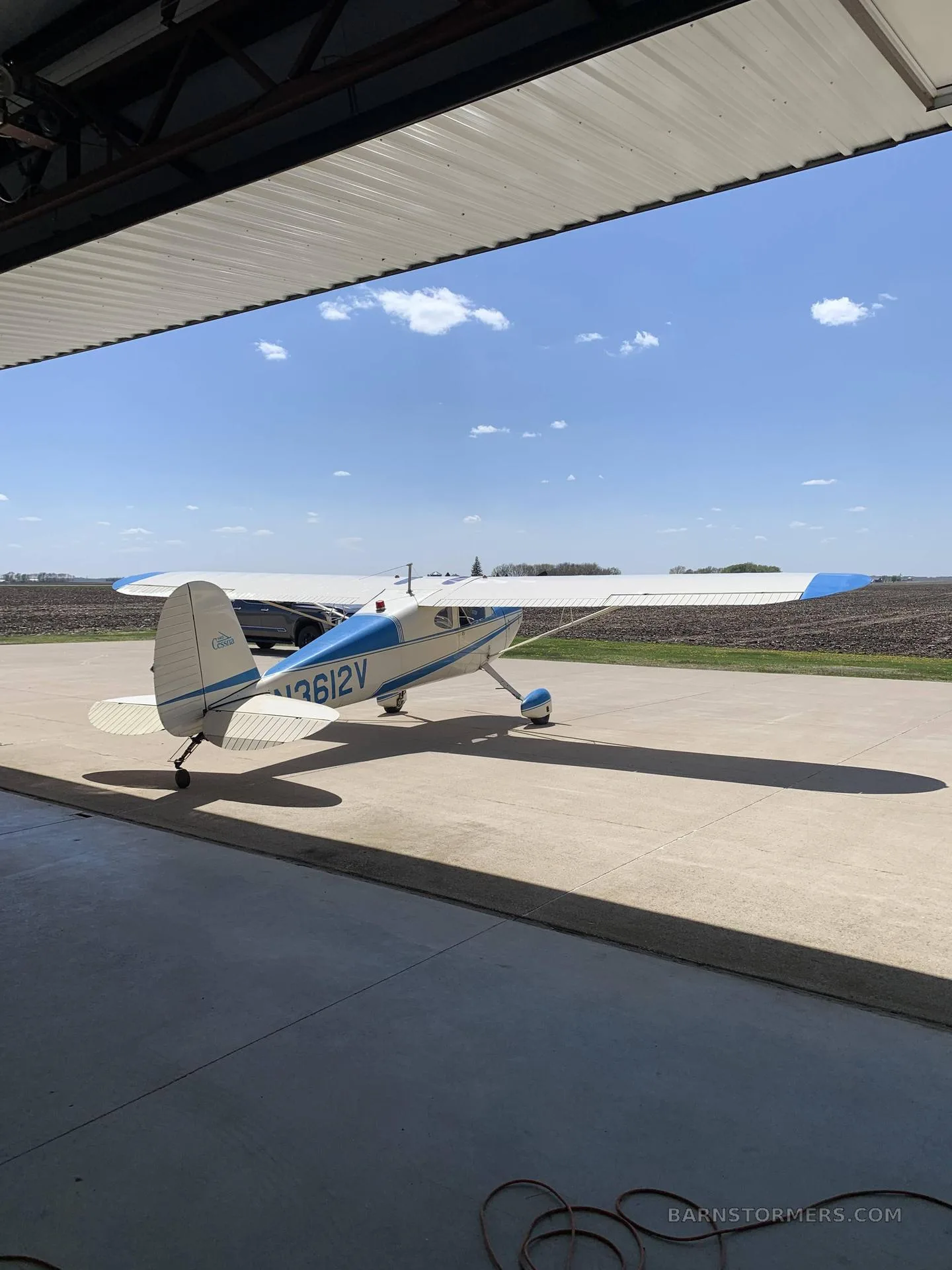 A white and blue Cessna 170B aircraft with tail number N3612V parked on a concrete pad, partially inside a hangar, with fields in the background.